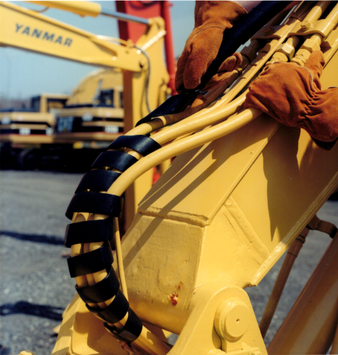 Close-up of a yellow excavator arm with black hydraulic hose wrap and gloved hands adjusting the linkage; YANMAR is visible in the background.